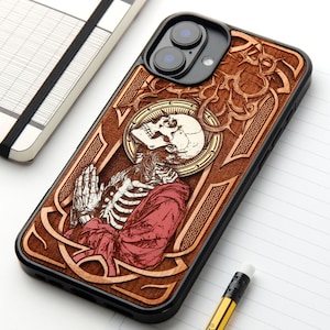 Könnte beinhalten: Eine schwarze Smartphone-Hülle mit einer holzfarbenen Einlage, die ein laser-graviertes Design eines Skeletts in roter Robe mit gefalteten Händen zeigt. Das Motiv umfasst einen kreisförmigen Heiligenschein hinter dem Schädel und verzierte Ränder.