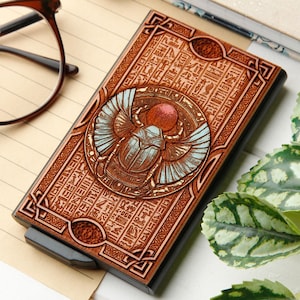 Könnte beinhalten: Ein Kartenhalter aus Holz mit einem ägyptischen Skarabäus-Design. Der braune Kartenhalter zeigt eine detaillierte Schnitzerei eines Skarabäus mit blauen Flügeln und einer roten Sonne, umgeben von Hieroglyphen. Der Kartenhalter ist rechteckig mit einem schwarzen Rahmen.