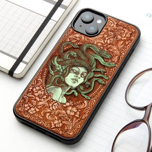 Può includere: Una cover per smartphone nera con un pannello posteriore in legno marrone finemente intagliato. Al centro, un'illustrazione in rilievo verde chiaro raffigura Medusa con serpenti al posto dei capelli, circondata da motivi floreali e volute.