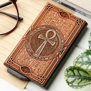 Könnte beinhalten: Ein Kartenhalter aus Holz mit einem gravierten Ankh-Symbol und Hieroglyphen. Der braune Halter hat einen schwarzen Rahmen und einen Druckknopfmechanismus. Das Ankh befindet sich in einem kreisförmigen Design mit Bronze-Finish. Der Kartenhalter liegt auf einer hellen Oberfläche.