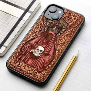 Op de afbeelding: Een zwarte smartphonehoes met een gedetailleerd in hout gegraveerd ontwerp van een figuur in gewaad die een kroon draagt en een menselijke schedel vasthoudt. De achtergrond is gevuld met complexe bloemmotieven en een pentagramsymbool.