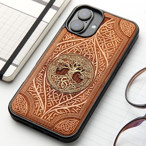 Op de afbeelding: Een zwarte smartphonehoes met een lichtbruine houten inleg en een ingewikkeld lasergegraveerd ontwerp. Het midden toont een cirkelvormig boommotief omringd door gedetailleerde Keltische knooppatronen over de gehele achterzijde.