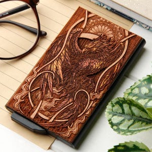 May include: A wooden card holder featuring a laser-etched phoenix design. The phoenix has detailed wings and feather patterns. The holder is brown with a black frame and a sliding mechanism. The dimensions are approximately 10 cm x 7 cm.