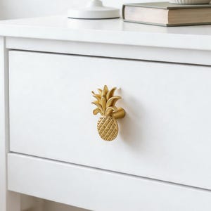 May include: A gold-colored pineapple-shaped drawer knob attached to the front of a white wooden nightstand. The knob features a textured, cross-hatched body and detailed leaves at the top, providing a functional handle for the drawer.