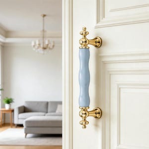 Pode incluir: Um puxador de porta vertical com uma secção central cilíndrica, lisa e azul-clara feita de cerâmica, acentuada por ferragens de metal em tom dourado polido na parte superior, inferior e nos pontos de fixação numa porta branca.