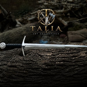 May include: A silver sword with a black handle and a decorative pommel rests on a textured, dark brown log. The sword is the focal point, with the logo "TALIA ART COLLECTIBLE" in gold above it.