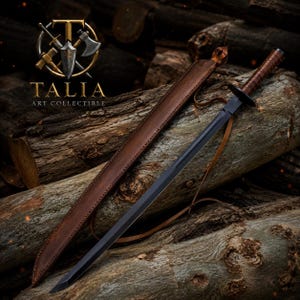 May include: A black sword with a brown leather-wrapped handle and a matching sheath. The sword is displayed on a pile of logs. The sheath has a strap. The text "TALIA ART COLLECTIBLE" is visible in the image.