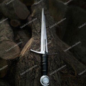 May include: A medieval sword with a silver blade and crossguard, a black handle, and a silver pommel. The sword rests on a textured, dark brown tree trunk, with a background of stacked logs. The sword is a collectible item.