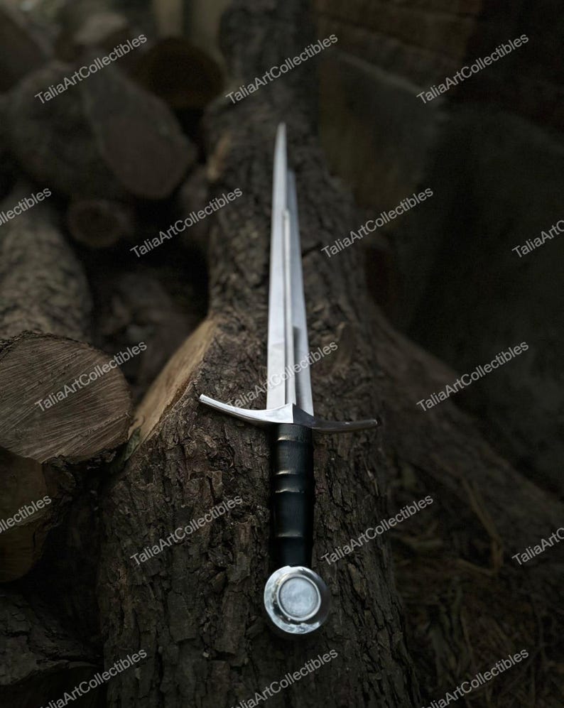 May include: A medieval sword with a silver blade and crossguard, a black handle, and a silver pommel. The sword rests on a textured, dark brown tree trunk, with a background of stacked logs. The sword is a collectible item.