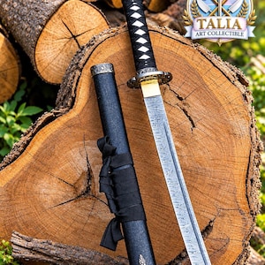 Op de afbeelding: Een katana zwaard met een stalen lemmet met patroon, een zwart omwikkeld handvat en een goudkleurige kraag. Het zwaard bevat een zwarte schede met een zwart lint en decoratieve metalen uiteinden. Een rond logo met de tekst TALIA ART COLLECTIBLE.