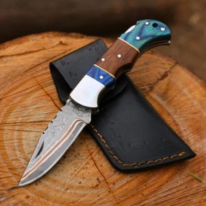 May include: A folding knife with a patterned blade and a wooden handle with blue and white accents. The knife is resting on a wooden surface next to a black leather sheath.