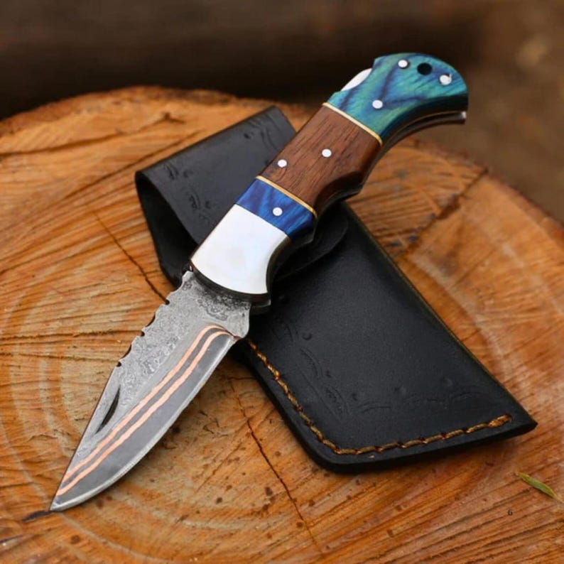 May include: A folding knife with a patterned blade and a wooden handle with blue and white accents. The knife is resting on a wooden surface next to a black leather sheath.