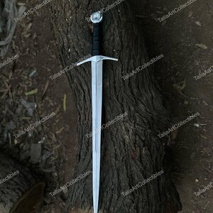 May include: A long, silver sword with a black handle and a cross-shaped guard. The sword has a pointed tip and a decorative pommel. The blade is made of polished metal. The sword is resting on a textured, brown tree trunk.