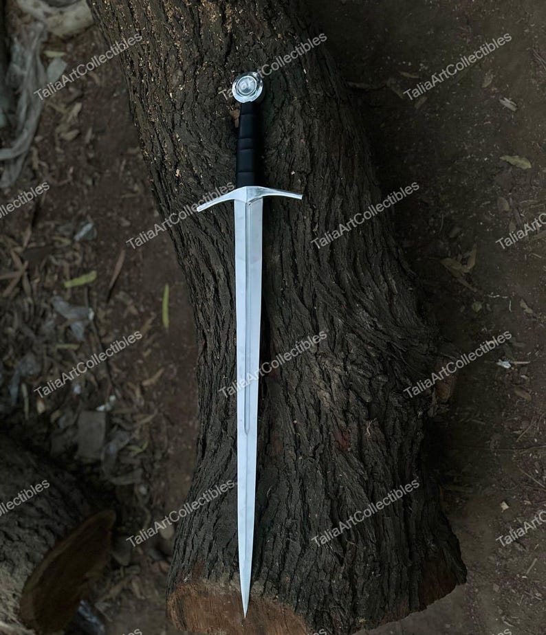 May include: A long, silver sword with a black handle and a cross-shaped guard. The sword has a pointed tip and a decorative pommel. The blade is made of polished metal. The sword is resting on a textured, brown tree trunk.