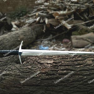 May include: A silver and black sword with a crossguard and a rounded pommel, resting on a textured, brown log. The blade is polished and reflects light. The sword's handle is black, and the crossguard is silver.