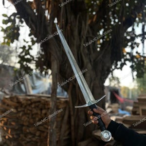 May include: A long, silver sword with a cross-shaped hilt and a black handle. The sword is held against a backdrop of a tree and a pile of wood. The text "TaliaArtCollectibles" is watermarked on the image.