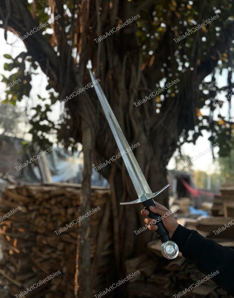 May include: A long, silver sword with a cross-shaped hilt and a black handle. The sword is held against a backdrop of a tree and a pile of wood. The text "TaliaArtCollectibles" is watermarked on the image.