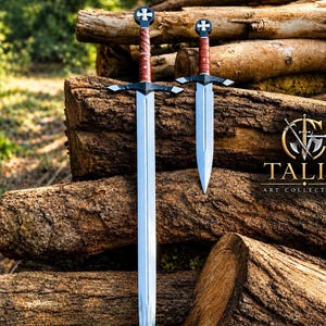 Peut inclure: Une épée longue et une dague plus courte, toutes deux dotées de lames argentées, de gardes noires et de poignées gainées de cuir marron. Chaque pommeau est surmonté d'une croix blanche. Le texte TALIA ART COLLECTIBLE est visible à droite.