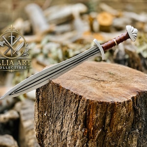 May include: A double-edged sword with a silver-colored blade featuring a central groove, a brown handle with a gold-colored ring, and a silver-colored pommel. The sword is marked with the text TALIA ART COLLECTIBLE and a logo.