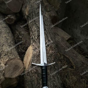 May include: A silver sword with a black handle and a crossguard. The sword is resting against a tree trunk. The blade is long and sharp, and the handle is wrapped in black material. The crossguard is silver and has a simple design.