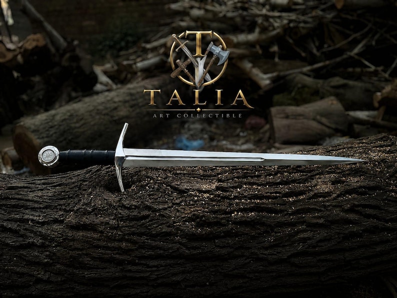 May include: A silver sword with a black handle and a decorative pommel rests on a textured, dark brown log. The sword is the focal point, with the logo "TALIA ART COLLECTIBLE" in gold above it.