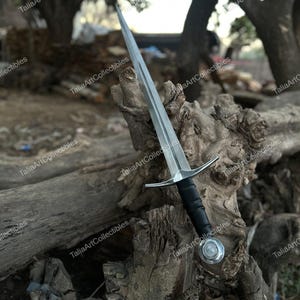 May include: A silver and black sword with a long, pointed blade. The sword has a black handle and a silver crossguard. The pommel is silver with a rounded design. The sword is displayed on a weathered wooden surface.