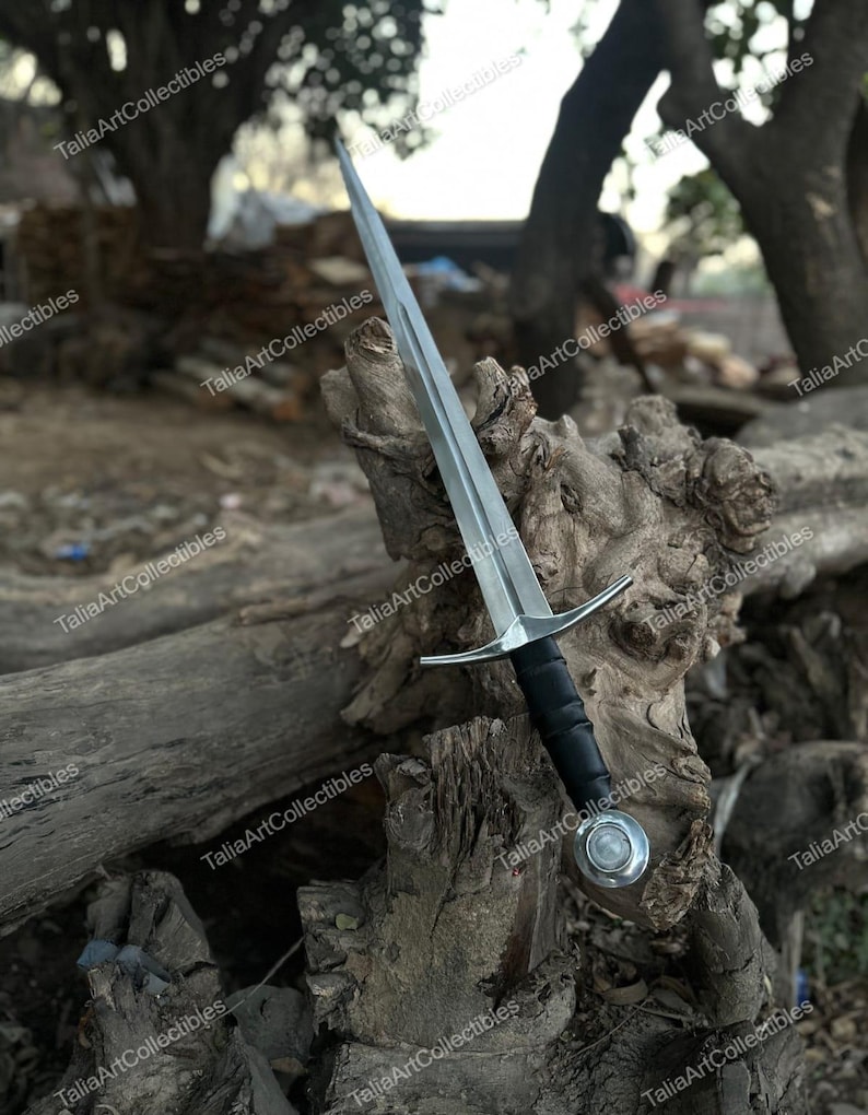 May include: A silver and black sword with a long, pointed blade. The sword has a black handle and a silver crossguard. The pommel is silver with a rounded design. The sword is displayed on a weathered wooden surface.