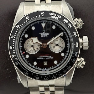 May include: A stainless steel Tudor Geneve chronograph watch with a black dial, two silver sub-dials, and a black tachymeter bezel. The watch features a date window at the six o'clock position, luminous hour markers, and a metal link bracelet.
