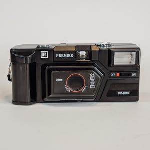 Może przedstawiać: Czarny aparat analogowy Premier PC-600 35mm focus free. Z przodu znajduje się prostokątna obudowa obiektywu z pomarańczowym tekstem, wbudowana lampa błyskowa po prawej stronie oraz przełącznik zasilania z oznaczeniami OFF i ON. Nazwa marki Premier jest nadrukowana na biało.