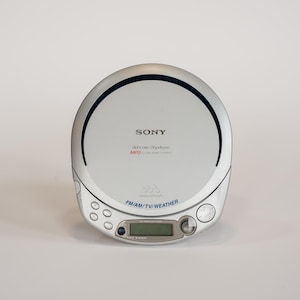 Può includere: Lettore CD portatile Sony argentato con display digitale e radio FM/AM/TV/Meteo. Il dispositivo ha un design circolare con una linea nera sulla parte superiore. Il testo "SONY" è stampato.