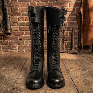 Puede incluir: Un par de botas negras hasta la rodilla con diseño de cordones frontales, ojales metálicos y un puño ancho en la parte superior asegurado por dos correas ajustables con hebillas. Las botas tienen punta redondeada y suela gruesa y resistente.