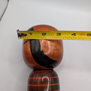 Op de afbeelding: Een houten kokeshi-pop met een ronde kop en cilindervormig lichaam, voorzien van een natuurlijke houtnerfafwerking, zwarte geschilderde gebogen lijnen en rode accenten. Een geel meetlint over de kop toont een diameter van 8,9 cm.