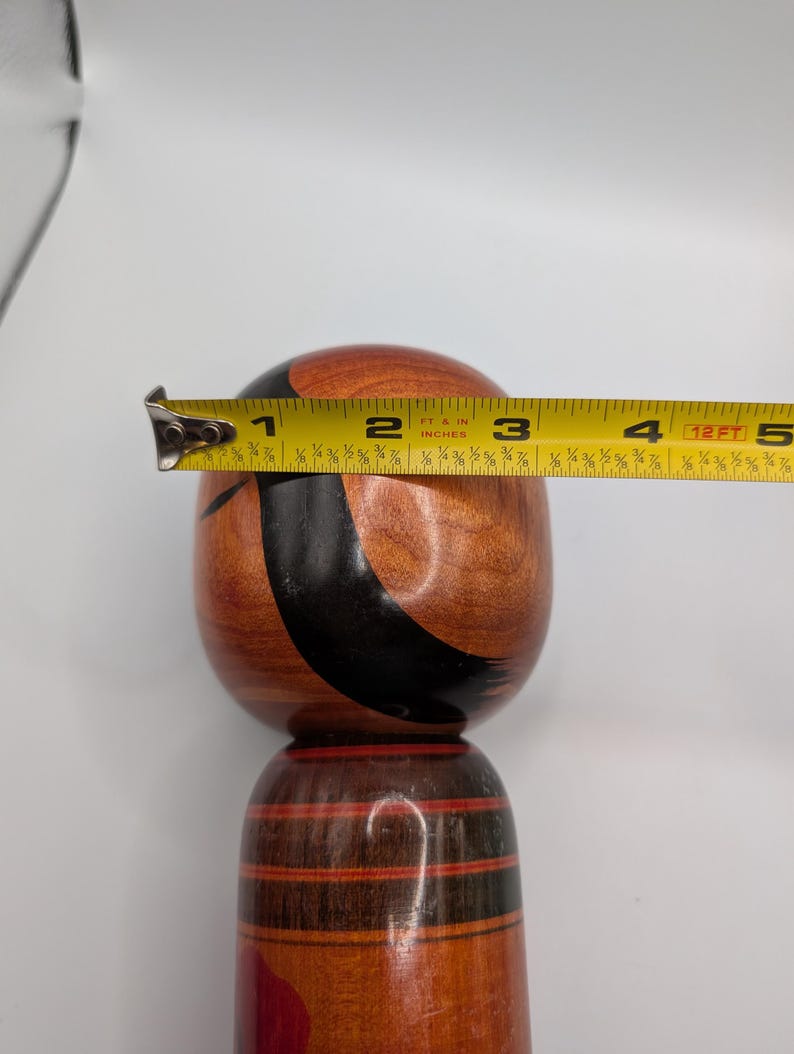 Op de afbeelding: Een houten kokeshi-pop met een ronde kop en cilindervormig lichaam, voorzien van een natuurlijke houtnerfafwerking, zwarte geschilderde gebogen lijnen en rode accenten. Een geel meetlint over de kop toont een diameter van 8,9 cm.