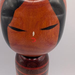 Op de afbeelding: Een houten kokeshi-pop met een glad, rond hoofd, zwart geschilderd haar, gesloten ogen, een kleine witte neus en rode lippen. Het cilindrische lichaam is afgewerkt in een roodbruine houttint met donkere horizontale banden en een bloemmotief.