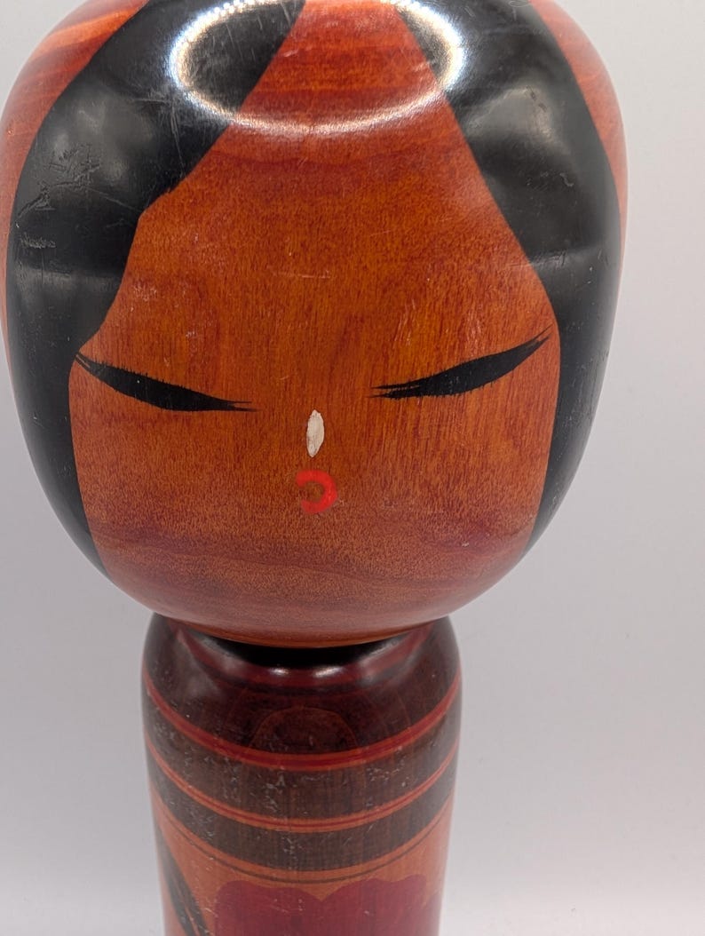 Op de afbeelding: Een houten kokeshi-pop met een glad, rond hoofd, zwart geschilderd haar, gesloten ogen, een kleine witte neus en rode lippen. Het cilindrische lichaam is afgewerkt in een roodbruine houttint met donkere horizontale banden en een bloemmotief.