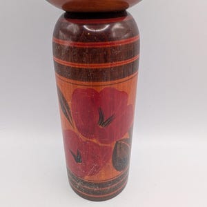 Op de afbeelding: Een cilindrische houten kokeshi-pop met een glad, rond hoofd en een taps toelopend lichaam. Het lichaam is afgewerkt in een warme bruine houttint met horizontale banden en een handgeschilderd bloemmotief in rood- en donkerbruintinten.