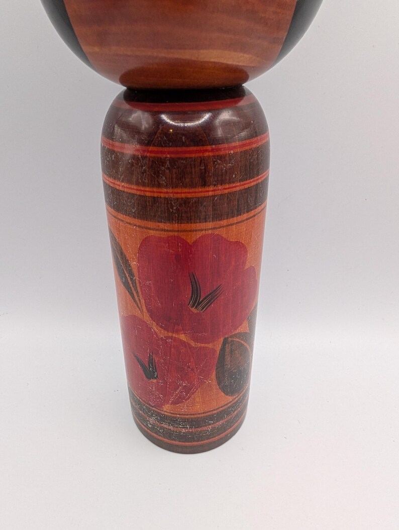 Op de afbeelding: Een cilindrische houten kokeshi-pop met een glad, rond hoofd en een taps toelopend lichaam. Het lichaam is afgewerkt in een warme bruine houttint met horizontale banden en een handgeschilderd bloemmotief in rood- en donkerbruintinten.