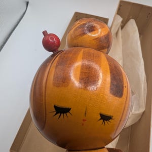 Op de afbeelding: Een vintage Japanse Kokeshi-pop met een ronde kop en lichaam. De kop van de pop is gestreept met tinten bruin en oranje, met gesloten ogen en een kleine rode mond. Een klein rood ornament zit bovenop het hoofd. De pop is gedeeltelijk zichtbaar in een kartonnen doos.