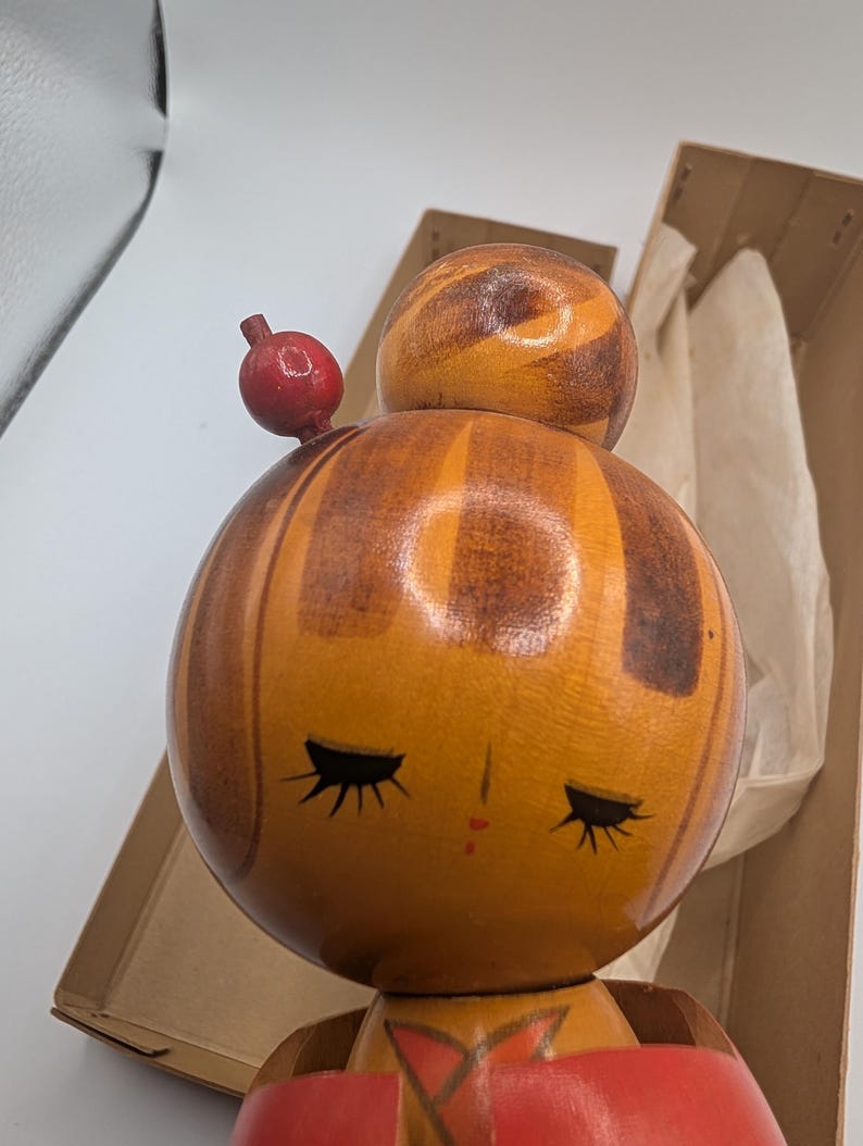 Op de afbeelding: Een vintage Japanse Kokeshi-pop met een ronde kop en lichaam. De kop van de pop is gestreept met tinten bruin en oranje, met gesloten ogen en een kleine rode mond. Een klein rood ornament zit bovenop het hoofd. De pop is gedeeltelijk zichtbaar in een kartonnen doos.
