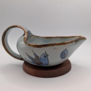May include: A light blue ceramic gravy boat with a single handle and a pointed spout. The exterior features a hand-painted blue bird motif with brown accents. The rim and handle edges are finished with a dark brown glaze, resting on a circular base.