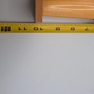 Op de afbeelding: Een houten deegroller met een rode handgreep ligt naast een gele meetlint. Het houten gedeelte van de deegroller meet ongeveer 25 cm.
