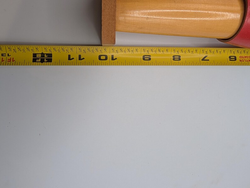 Op de afbeelding: Een houten deegroller met een rode handgreep ligt naast een gele meetlint. Het houten gedeelte van de deegroller meet ongeveer 25 cm.