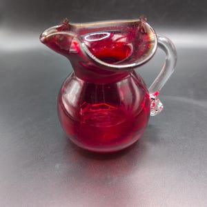 Op de afbeelding: Een kleine, dieprode glazen kan met een rond lichaam, een brede, gegolfde schenktuit en een helder, gebogen glazen handvat aan de zijkant. Het doorschijnende rode glas heeft een gladde, gepolijste afwerking met een levendige kleur.