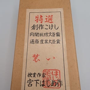 Op de afbeelding: Een rechthoekige kartonnen doos met een cr&egrave;mekleurig etiket met Japanse tekst in rode en zwarte inkt. De tekst bevat "特選" en "創作こけし". Het etiket heeft ook een rode zegel en de naam "宮下はじめ作".