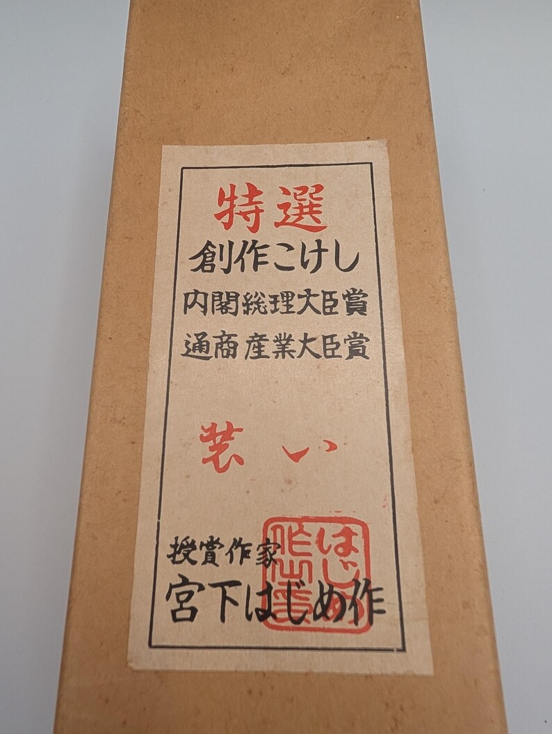 Op de afbeelding: Een rechthoekige kartonnen doos met een cr&egrave;mekleurig etiket met Japanse tekst in rode en zwarte inkt. De tekst bevat "特選" en "創作こけし". Het etiket heeft ook een rode zegel en de naam "宮下はじめ作".