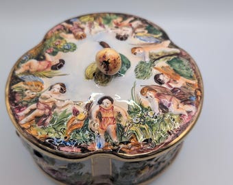 May include: A round porcelain trinket box with a scalloped edge and a gold-trimmed lid. The lid features a colorful painted scene of multiple cherubs playing in a grassy landscape, topped with a small, round, patterned knob handle.
