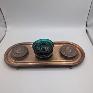 May include: An oval copper desk tray featuring a central teal glass inkwell and two circular copper lidded containers on either side. The tray has a raised rim and small feet, with decorative floral patterns embossed on the lids of the containers.