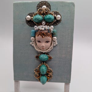 May include: A vintage brooch featuring a face, turquoise stones, and faux pearls. The brooch is mounted on a pale blue canvas, with a gold-toned filigree design. The face has painted details, and the brooch is displayed on a small easel.