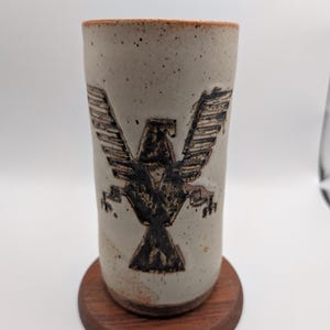 Op de afbeelding: Een cilindervormige keramische beker met een gespikkeld gebroken wit glazuur en een donkerbruine, gestempelde adelaarsafbeelding op de voorzijde. De beker heeft een ongeglazuurde rand en voet in terracotta-kleur en staat op een ronde onderzetter van donker hout.