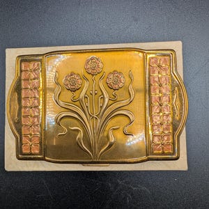 May include: A rectangular brass belt buckle featuring a central embossed floral design with three flowers and long stems. The side panels are decorated with a grid of small, pink-enameled flower motifs. The metal has a polished, golden finish.